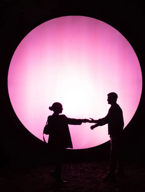 Stefano Porta/LaPresse/Shutterstock Two people are in silhouette in front of a a large pink circle, light display  