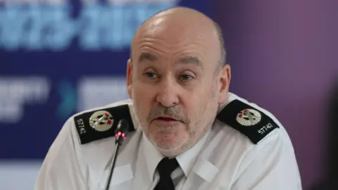 PA Media PSNI ACC Ryan Henderson, a man with grey hair at the sides and grey stubble. He has a white shirt with black lapels and a black tie.