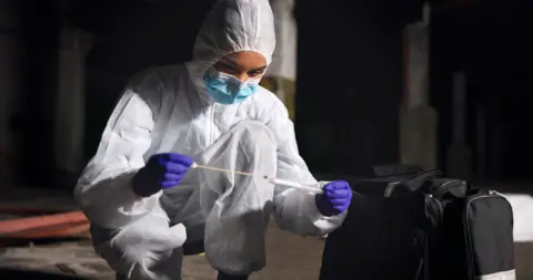 Getty Images Generic stock image of a forensic scientist working at a crime scene. It is dark, she wears a white hazmat suit with the hood up, a face mask on and purple rubber gloves, as she takes a sample from the scene and puts it into her test tube.