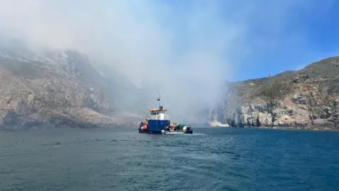 Andy Cook. Un barco en el mar azul. Detrás del barco está Little Sark. Sale humo gris claro.