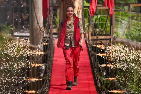 Shutterstock Tulisa Constostavlos walks across the I'm A Celebrity bridge wearing a beige shirt, red gilet and red trousers as fireworks go off around her.