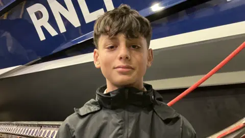 Kaan, a boy who is wearing a black waterproof coat. He is standing in front of a blue RNLI boat.