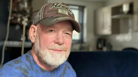 BBC Emmanuel Burns has a short white beard and is wearing a brown baseball cap and a blue t-shirt. He is sitting inside his apartment.