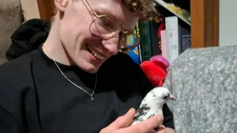 Jack Chadwick Jack Chadwick, who is wearing a black T-shirt, is sitting on a couch as he smiles and holds the grey and white pigeon.
