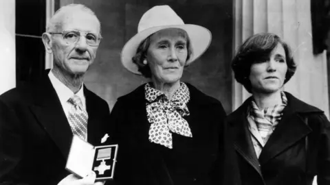 Getty Images/Central Press Archive image showing Nairac's family, father Maurice with short gray hair and glasses, mother Barbara with a white hat and scarf and sister Rosamonde with short black hair and a dark jacket