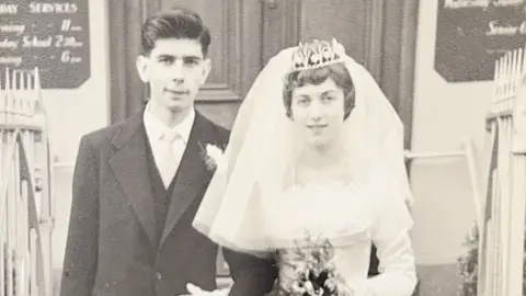 A man and a woman are celebrating their wedding day. The woman is wearing a wedding dress with a veil and tiara, and the man is wearing a black suit with a white shirt. The picture is in black and white. 
