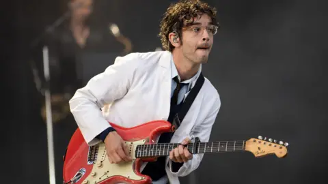 PA Media La Matty Healy de 1975, vestida con un traje blanco, tira de la cara mientras toca la guitarra en el escenario en un festival en Escocia, 2024