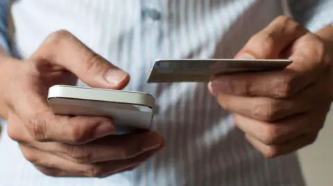 Getty Images Stock close-up image of a man holding a phone in his right hand and a bank card in the other. He is wearing a blue-and-white striped shirt.