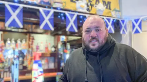 Liam Logue - a man wearing a dark hoodie with Scotland flags and bunting at the bar behind him