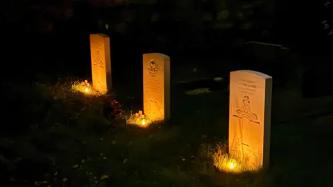 Home Front Museum, Llandudno A row of three white gravestones arranged in a diagonal row across the shot.  It's night time so the area around them is in darkness.  Each has a small battery night light in front of it, so they are lit up in a warm orange glow.