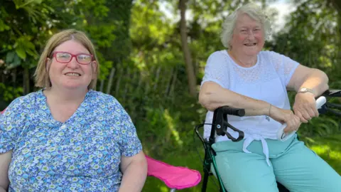 Two women sat facing the camera. The woman on the left is wearing a blue patterned top and has a bob and is wearing pink glasses. The woman on the right is wearing light blue trousers and a white top. Both are looking at the camera smiling. Behind then is green grass and trees.  