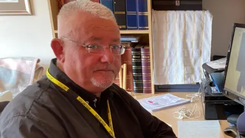 A close up of Darren Gilchrist. He has short grey hair and is wearing glasses. He is also wearing a black shirt.
