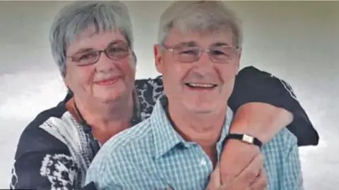 Sandra Francis Picture of a woman hugging a man from behind. Both couple have grey hair, are wearing glasses and smiling. The man is wearing a blue and white checked shirt while his wife is wearing a black and white shirt, a black chunky beaded necklace and a square framed watch with black leather straps.