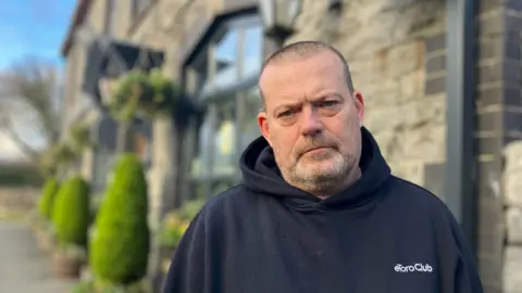 BBC Nils Kirk is standing outside the pub, wearing a dark coloured hooded top. He looks concerned. The stone-built pub with plants and shrubs in pots can be seen behind him.