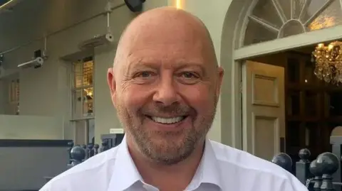 Craig Dowie looks at the camera. He has a bald head and light brown stubble. He is wearing a white shirt.