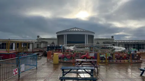 George Carden/BBC The area where the pool used to be which is now filled with children's amusement rides. To the left are trampolines, a mini-roller coaster in the middle and the bandstand can be seen behind. The rides are all closed because it is winter season