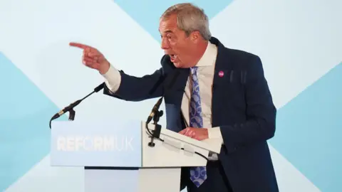 PA Media Nigel Farage pointing from a podium in front of a white and blue background.