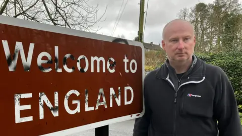 Roland Underdown is wearing a black fleece and is standing by a brown road sign which says Welcome to England.