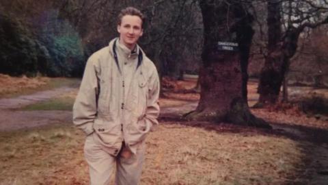 Trevor O'Donnell stands in a beige jacket in woodland in front of a large tree