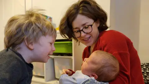 Jonny Coxon Charlotte in a red jumper holding a baby with a toddler next to her
