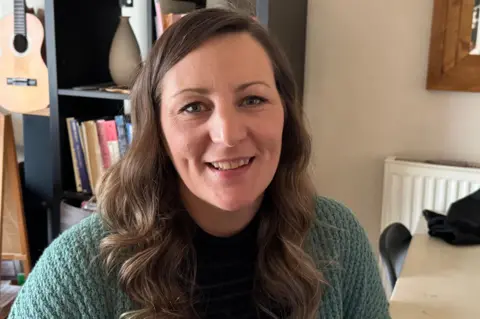 A woman wearing a teal green cardigan with long brown hair looks directly into the camera and smiles. There is a black book case and guitar in the background.