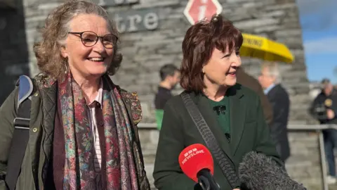 Marella Fyff has long, curly grey/brown hair and glasses while Fidema O'Kane has dark brown/red hair/ Both are standing in front of microphones 