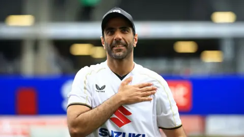 MK Dons Fahad Al Ghanim with his hand on his heart, wearing a baseball cat, football top, he has a beard and is smiling.