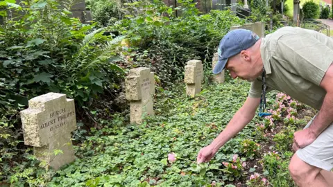 A man wearing a green shirt, light-coloured shorts, blue cap, and grey shoes is bending over to tend to flowers in a cemetery. The cemetery contains gravestones of various shapes and sizes, some covered in moss and surrounded by greenery. A pathway runs through the area, with trees and bushes in the background. One gravestone near the man reads 'HIER RUHT AUGUSTE PHILIPP.