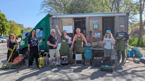 A group of people from Share Shed stand outside the Share Shed van. They are wearing green aprons and holding various items of electrical equipment.