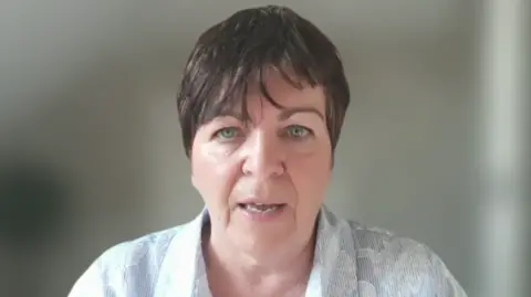 I woman with dark hair is speaking directly to the camera. She has short dark hair and is wearing a pale blue and white shirt with applique flowers. 
