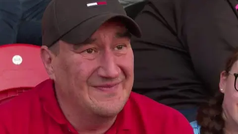 Ady Good wearing a black cap and a red polo shirt is sat in a crowd at a stadium