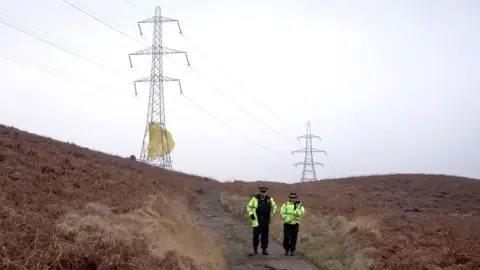 PA Media A male and a female police officer walk down a remote dirt track, which is flanked on both side by gorse. On the horizon are two large electricity pylons, there is yellow-coloured material that is wrapped around the bottom of the nearest pylon that appears to be billowing in the wind 