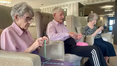 Two women and one man sit in armchairs knitting. The woman to the left has grey hair, glasses, a pastel pink blouse, a purple skirt and is knitting using green wool. The man sat in the middle has grey hair, a pink shirt and black tracksuit bottoms. He is knitting using bright pink wool. To the right is a woman in a black and white speckled blouse and a dark skirt. She also has grey hair and is knitting using white wool.