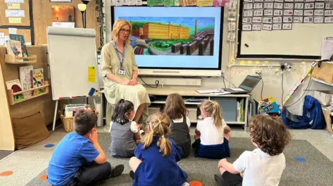 Aisha Iqbal/BBC A classroom scene. A teacher wearing a green and white floral dress sits at the head, and six children sit in front of her on the floor with their backs to the camera. Behind the teacher is a large piece of artwork on a screen, depicting a mill building and rows of houses in front of it.
