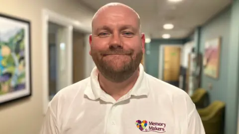 A man with a closely shaved head and a beard is smiling at the camera. He is wearing a white polo shirt with a Memory Makers logo on it