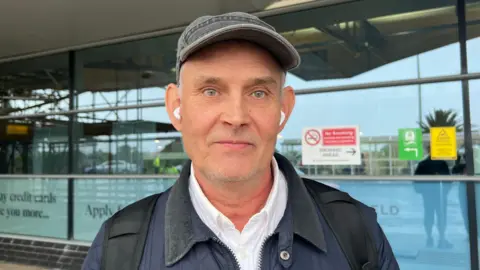 James Smith pictured wearing a grey cap and earbuds standing in front of an airport building. He is wearing a navy quilted jacket.