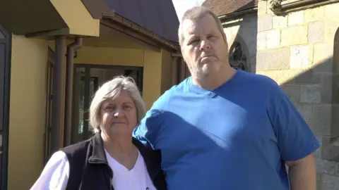 LDRS A man with short grey hair wearing a blue t-shirt. He is stood beside a woman with short grey hair wearing a pink t-shirt and black sleeveless jacket. They are looking into the camera and are stood outside of a yellow building