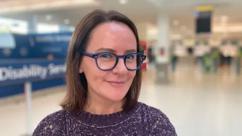 A young woman wearing a purple sweater, blue glasses and short hair is stood in an airport