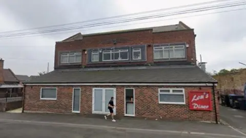 Google Streetview of a two-storey brick building, with a cafe downstairs with a large red sign reading "Lou's Diner" on the wall, and windows upstairs covered with metal bars.