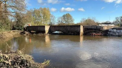 A low brick bridge over a river is pictured with trees at one end and scaffolding and sand bags at the other where it has been damaged. The underside of the bridge is a series of arches. 