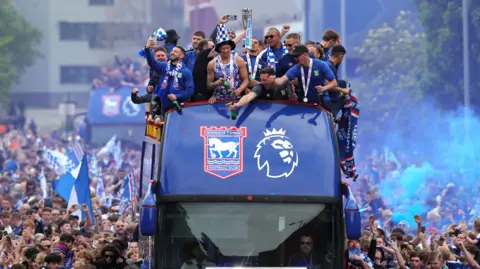 Blue Ipswich bus with players on top celebrating. There is a large crowd surrounding the bus and blue smoke in the air from flares.