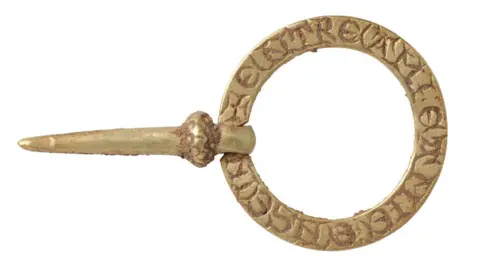 A 13th or 14th Century brooch, made of a circular band of gold, showing the inscribed back with mud on it, and a pin attached on one side of the band with a loop.