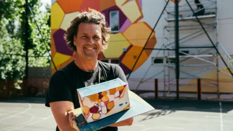 Helen Murray Patricio Forrester holding a model draft of the mural behind him, which features red, orange and yellow colours.