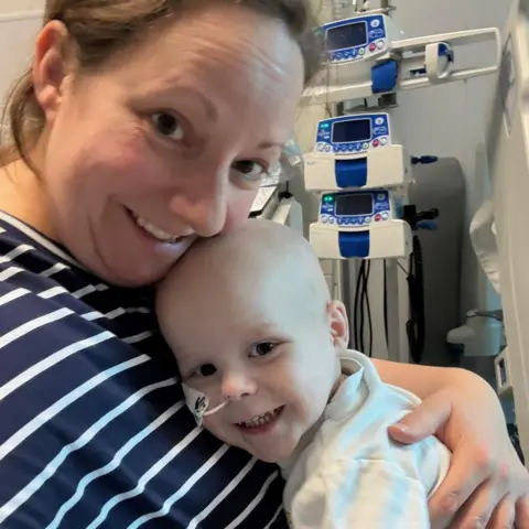 Sarah Sloman A woman with tied-back brown hair wearing a navy and white striped top. She is hugging a young boy wearing a white t shirt, who has a feeding tube in his nose. They are sat with hospital equipment behind them and are both looking at the camera and smiling.