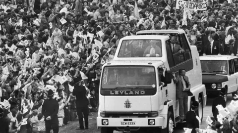 Getty Images Black and white image of an open-top white Leyland truck carrying the Pope and officials past a huge crowd of people waving at Heaton Park