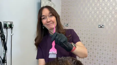 Lisa Crawford, who is using a pink brush to lay a product on a customers' head, is looking up and smiling slightly at the camera. She is wearing a purple, short-sleeved uniform top and has long brown hair. Hair straighteners are plugged in the wall behind her.