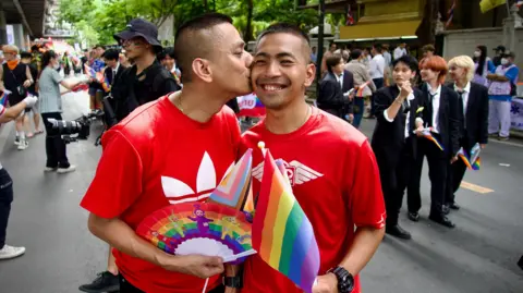 Benjamin Begley/ BBC Chanatip "Jane" Sirihirunchai kisses his partner Pisit "Kew" Sirihirunchai on the cheek on a Bangkok street during a Pride celebration. They are smiling in red shirts and wearing rainbow flags.  