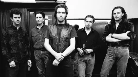 Getty Images Black and white photo of Del Amitri. They are standing in a hallway and are all wearing jeans and shirts. Justin Currie is also wearing a leather waistcoat
