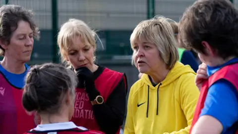 Simon Thurlow Janette Hynes has short blonde hair and a yellow sports top. She is standing with four women who have brown hair. Two have blue tops and one has a dark top, the three of them have red bibs on. They are on a football pitch surrounded by a green cage. They are receiving coaching instructions from Janette Hynes.