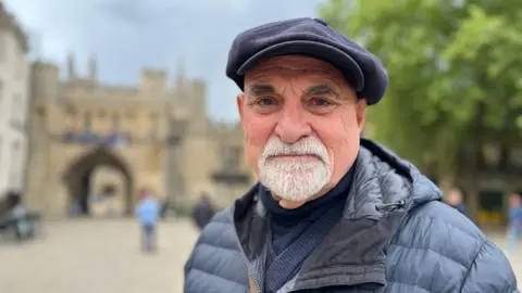Emma Baugh/BBC A man with a grey beard wearing a navy blue puffa jacket and a cap standing in front of a cathedral arch, with shoppers in the background.